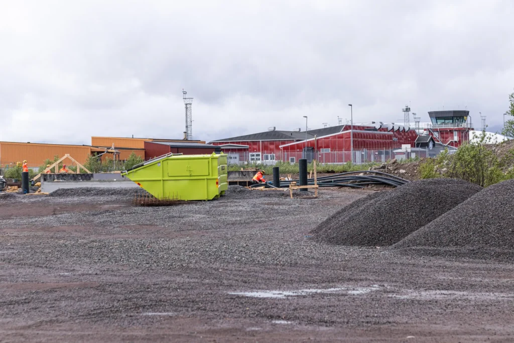 Keskeneräinen rakennustyömaa, jossa on soraa, vihreä siirtolava ja rakennusmateriaaleja. Taustalla näkyy Kiirunan lentoaseman punainen rakennus ja lennonjohtotorni.